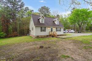 A small, beige, two-story house with a dark roof is surrounded by a grassy yard and trees. The house has dormer windows and a small porch with stairs leading to the front door. A gravel driveway on the right has parked vehicles.