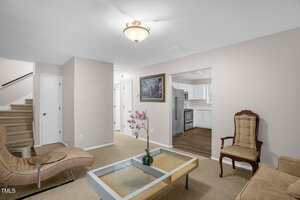 A living room with beige walls and carpet, featuring a glass coffee table with a pink flower arrangement. There's a cushioned chair, a modern recliner, and a view into a kitchen with white cabinets and wooden flooring. A staircase is visible to the left.