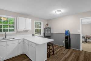 A kitchen with white cabinets and countertops, featuring a sink, under a ceiling light. There's a window above the counter. A water cooler stands to the right, near a small cabinet, with an entrance to another room visible in the background.