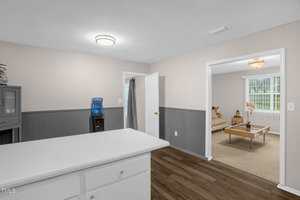 Kitchen with white cabinets and a countertop, leading to a living room with a beige sofa and wood table. Walls are adorned with wainscoting, and a water cooler is near the door. A light fixture is on the ceiling, and the flooring is wood-style.