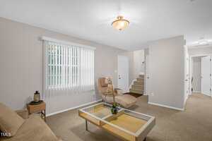 A living room with beige walls and carpet, featuring a brown sofa, a glass coffee table, and a tan cushioned chair with a footrest. A window with vertical blinds is on the left, and a staircase with white railing is visible in the background.