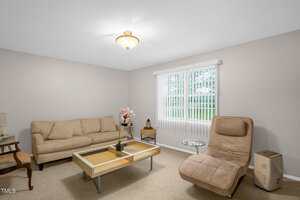 Living room with beige walls and carpet. Features a beige sofa, matching lounge chair, glass-top coffee table, small side table with a plant, and window with vertical blinds. Ceiling light fixture and a wooden chair in the corner.