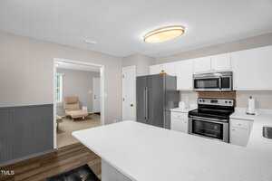 A kitchen with white cabinets, a silver refrigerator, and a stainless steel oven and microwave. A large white countertop is in the foreground. An adjoining room with beige walls and carpet is visible, furnished with a beige chair and a coffee table.