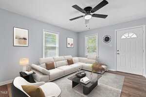 A modern living room with light blue walls, featuring a white sectional sofa and a white armchair. There's a black glass coffee table, two paintings, a clock on the wall, and a ceiling fan. Natural light enters through two windows with blinds.