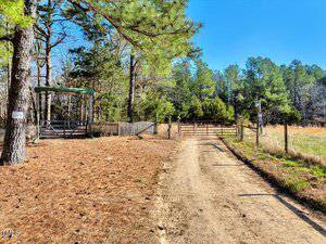A dirt road leads to a gated entrance in a rural area. The path is lined with trees and a wooden fence. Signs are attached to the gate and nearby fencing. The sky is clear, and the area is surrounded by greenery.