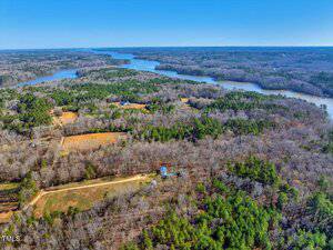 Aerial view of a expansive landscape showing dense forests with bare trees, a few open clearings, and a winding river or lake on the horizon. A dirt road runs through the scene. The sky is clear and blue.