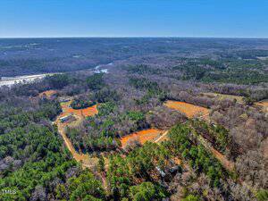 Aerial view of a vast rural landscape with dense forests, open fields, and scattered buildings. A winding dirt road cuts through the trees. A large body of water is visible in the distance under a clear blue sky.