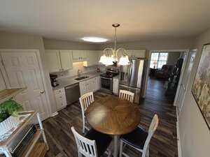 Spacious kitchen with white cabinets, stainless steel appliances, and a wood-topped round table with four chairs. Pendant light hangs above the table. Hardwood flooring and adjacent living area with a couch are visible. Large windows provide natural light.
