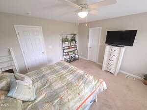 Bedroom with beige walls and carpet, featuring a bed with a floral comforter, a tall white dresser with a TV on top, a ceiling fan, a metal shelf with plants, and two closed white doors.
