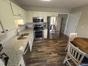 A kitchen with white cabinets, granite countertops, and stainless steel appliances including a microwave, stove, and refrigerator. There is a round wooden table with two chairs on a hardwood floor.