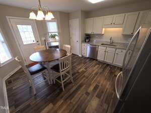 A kitchen with wood-patterned flooring, white cabinets, and stainless steel appliances. A round table with four wooden chairs sits in the center. A door and window are on the left, and a chandelier hangs above the table.
