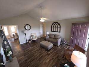 Living room with wood flooring, a sofa, ottoman, and a rocking chair. A ceiling fan hangs from the vaulted ceiling. A round mirror and window-shaped decor adorn the walls. A dining area is visible through an open doorway.