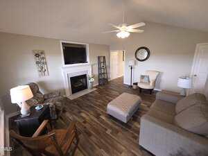 A cozy living room features a ceiling fan, a gray sofa, and patterned armchairs. A flat-screen TV is mounted above a white fireplace. The wood floor is complemented by a large ottoman and a round wall mirror. Decorative plants are placed beside the fireplace.