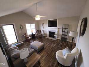 A cozy living room with hardwood floors features a sofa, an armchair, and a recliner chair. A fireplace topped with a TV is centered on the far wall. Near the window are a floor lamp and a small table. A ceiling fan with a light is above.