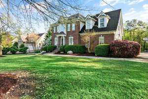 A two-story brick house with white trim and a black shingle roof sits amid a neatly landscaped yard. The front lawn is expansive with trimmed bushes lining the pathway. A basketball hoop is visible on the right side of the driveway.