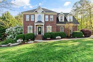 A two-story brick house at 1154 Smith Creek Way in Wake Forest boasts a manicured lawn and neatly trimmed shrubs. The front features five tall windows, an ornate glass door centered with steps leading up, and is surrounded by trees and bushes under a partly cloudy sky.