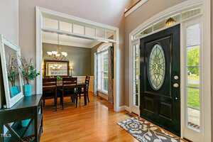 Entryway with a black door featuring decorative glass. Adjacent is a dining room with a dark wood table and chairs, a mirror, and a chandelier. A side table with turquoise vases is on the left. Light streams through a large window with curtains.