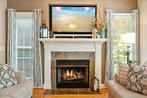 A cozy living room features a lit fireplace with a white mantel. Above, a TV displays a beach scene. The mantel is decorated with vases and lanterns. Two windows with patterned curtains flank the fireplace, and there is a cushioned chair on each side.