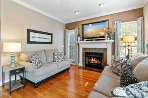 A cozy living room with wooden flooring features two beige sofas, a framed picture, and a lit fireplace with a TV above it. Lamps are placed beside each sofa, and large windows provide natural light.