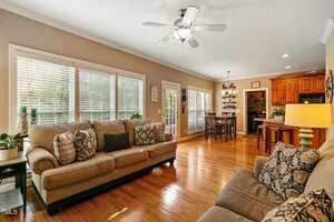 A living room with two sofas, a ceiling fan, and hardwood floors. Large windows and a glass door let in natural light. The space opens to a dining area with a wooden table and chairs, and a kitchen with wooden cabinets and an island.