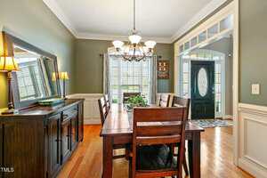 A dining room with a wooden table and six chairs is shown. A large mirror hangs above a sideboard on the left. A chandelier is centered over the table. The room has green walls and a hardwood floor. A door with stained glass is visible in the entryway.