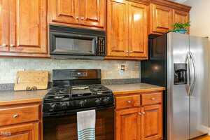 A kitchen featuring wooden cabinets, a stainless steel refrigerator with a water dispenser, and a black stove and microwave. A cutting board and striped towel are visible, with a light tiles backsplash completing the look.