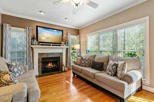A cozy living room with light beige walls and wooden floors. There's a fireplace with a TV above it. Two sofas with patterned cushions face the fireplace. Large windows let in natural light, and a ceiling fan is overhead.