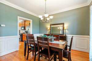 A dining room with a dark wooden table and six matching chairs. There's a hanging chandelier above the table. The walls are painted green with white wainscoting. A mirror and two lamps are on a sideboard, and a kitchen is visible through the doorway.