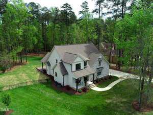 Aerial view of a two-story white house with a grey roof, surrounded by a large green lawn and trees. The property includes a driveway and a fenced backyard. The house features a covered front porch and several windows.