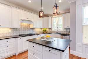 A modern kitchen with white cabinets, black countertops, and a central island. The backsplash features a patterned design. Two pendant lights hang above the island, and a bowl of lemons is placed on it. A window above the sink lets in natural light.