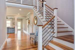 A modern home interior featuring a wooden staircase with metal railings. The hallway has hardwood floors, a wall mirror, and a small bench with cushions. A doorway leads to a living area with a couch and large windows.