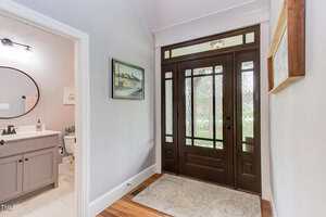A hallway featuring a dark wooden front door with frosted glass panels. To the left is a doorway leading to a bathroom with a vanity and toilet visible. The walls are light-colored, and a framed artwork hangs above the door.