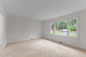 An empty living room with light beige carpet and off-white walls. A large window on the right wall overlooks a residential street with trees and a parked car. The room is well-lit with natural light.