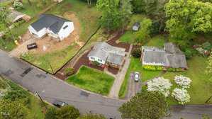 Aerial view of a suburban area featuring two houses with yards and driveways, a white car parked in one driveway, a small shed in a garden area, and a newly constructed building with soil around it. A road divides the properties.