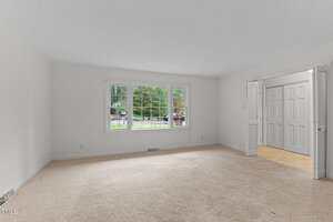 An empty living room with white walls and beige carpet. A large window on the left wall shows a view of greenery outside. On the right, there is an open doorway leading to another room or hallway.
