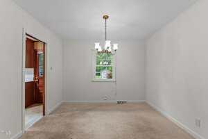 A carpeted dining room with white walls and a central chandelier. There is a window on the far wall showing greenery outside. An open doorway leads to an adjacent room with visible wood paneling.