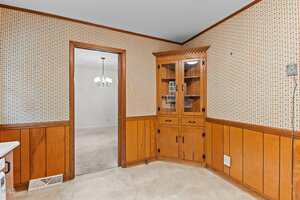 A kitchen corner features wood paneling and a built-in wooden cabinet with glass doors displaying small items. The beige walls have a patterned design. A doorway on the left leads to a carpeted room with a chandelier. The floor is tiled.
