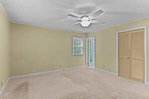 A spacious, empty bedroom with beige carpet, light yellow walls, a white ceiling fan, a window with blinds, a closet with louvered doors, and a doorway leading to a bathroom.