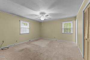 A spacious, empty bedroom with beige carpet, light yellow walls, two windows with white blinds, a ceiling fan with lights, and double closet doors. An electrical cord lies on the floor near the wall.