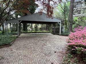 A brick-paved driveway leads to a carport surrounded by dense greenery and trees. Pink flowers bloom on bushes to the right, and a wooden fence is partially visible through the foliage.