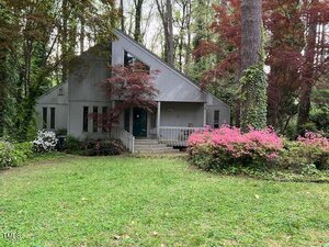 Nestled among tall trees at 106 Orchard Road, Henderson, this modern gray house features a striking triangular roof. The front boasts a cozy porch surrounded by pink and white flowering shrubs, while a lush grassy lawn stretches invitingly to the foreground.