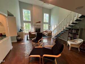 Spacious living room with high ceilings, large windows, and a white brick fireplace. There are two black chairs, a coffee table, and two sofas. A staircase leads to an upper floor. Bookshelves and decorative items are arranged neatly.