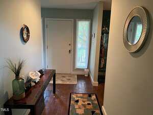 A hallway with brick flooring features a modern wooden bench on the left, decorated with a vase and small items. A round mirror hangs on the right wall. A door with a vertical window and a side window leads outside. Rugs are placed on the floor.