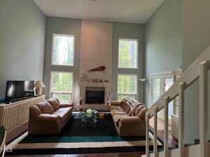 Spacious living room with high ceilings and large windows. Two brown leather sofas face a modern coffee table and a television. A brick fireplace is centered on the main wall, with decorative items on the mantel. A staircase is visible on the right.