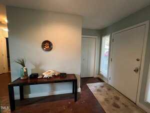 A hallway with a wooden table against a light blue wall. The table holds a vase with grass, a bowl, and other small items. A decorative plate hangs above the table. A closed white door and a window with blinds are on the right. A beige rug is on the floor.