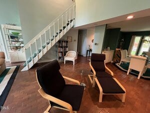 A living room with brick flooring features two black lounge chairs with wooden frames. A staircase with white railings leads upward. A small bookshelf and decorative items sit against the wall. A dining area is visible in the background.
