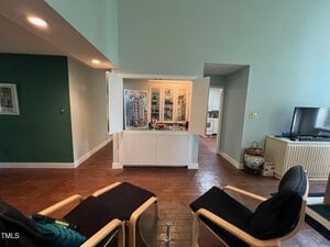 A room with light green walls, brick flooring, and two black chairs. A white cabinet is open, displaying various items inside. A basket and TV are on the right. A hallway leads to another room at the back.