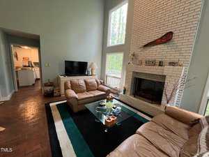 Living room with tall white brick fireplace, large windows, and brown leather sofas. A glass coffee table sits on a rug, and a flat-screen TV is near a hallway leading to a kitchen. Walls are light blue-gray, with a canoe paddle and fireplace decor.