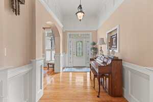A hallway with wooden floors, white wainscoting, and light-colored walls. A piano with a lampshade and framed artwork sits on the right. An arched opening leads to another room. A front door with decorative glass panels is visible at the end.