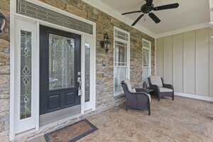 A porch with a stone wall features a black front door with decorative glass panels and two small windows. Two wicker chairs with cushions are placed on the tile floor. A ceiling fan hangs above, and wall-mounted lanterns flank the windows.